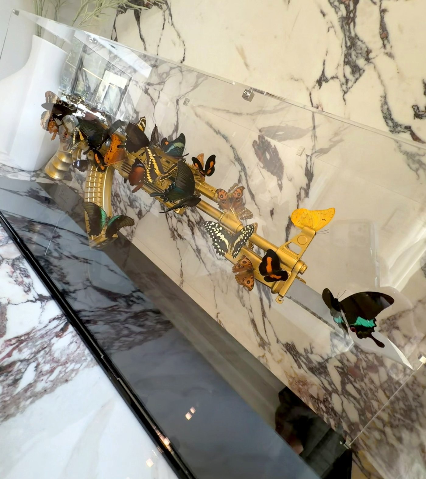 Gold rifle with bayonet and butterflies in a clear case against veined marble